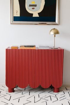 a red sideboard with books on it in front of a painting and a lamp