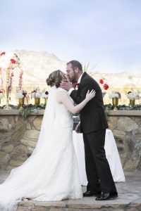 a bride and groom kissing in front of a stone wall with red flowers on it