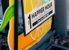 a sign that says happy hour on the side of a street with oranges and lemons