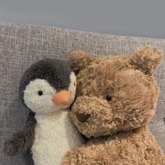 two stuffed animals sitting next to each other on a couch with one penguin hugging the other