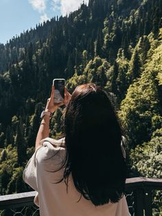 a woman taking a photo with her cell phone in front of a mountain range and forest