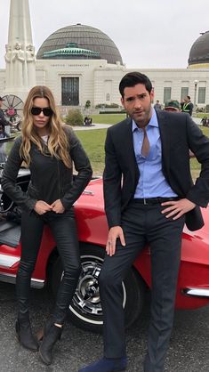 two people sitting on top of a red sports car in front of a large building