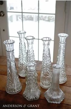 four glass vases and one candle holder on a wooden table in front of a window