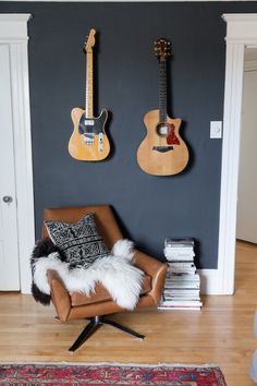 two guitars are hanging on the wall next to a leather chair and bookshelf