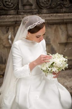 a woman in a white wedding dress holding a bouquet and looking down at the ground