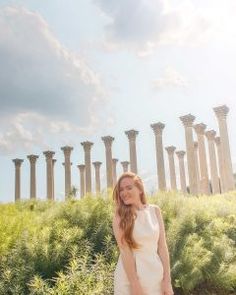 a woman in a white dress standing next to some tall pillars and grass on a sunny day