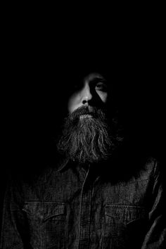 a man with a long beard standing in the dark wearing a denim shirt and jeans