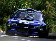 a blue car driving down a road next to some bushes and trees in the background