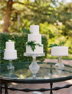 a table topped with three white cakes covered in green leafy decorations on top of glass pedestals