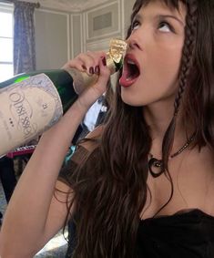 a woman in a black dress is drinking from a wine bottle and holding a cork to her mouth