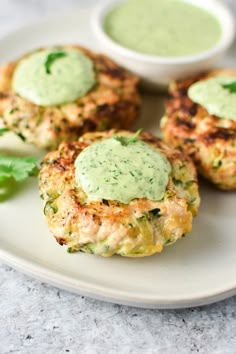 three crab cakes on a white plate with some green sauce in the middle and two small bowls next to them