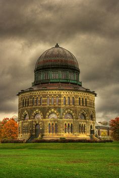 an old building with a dome on top in the middle of a grassy field under a cloudy sky