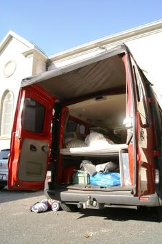 an open red van parked in front of a white building with its doors open and luggage piled on the floor