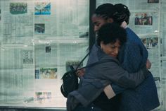 two women hugging each other in front of newspaper pages on the wall with posters behind them