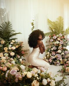 a woman sitting on the ground surrounded by flowers and greenery with her arms crossed