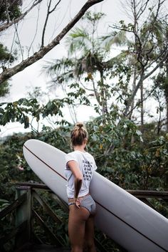 a woman holding a white surfboard on top of a wooden platform in front of trees