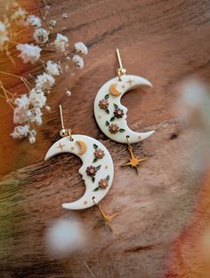 two white moon and star shaped earrings on top of a wooden table next to flowers