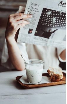 a person sitting at a table reading a newspaper and eating bread with a glass of milk