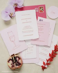 the wedding stationery is laid out on top of each other, including pink envelopes and wine glasses