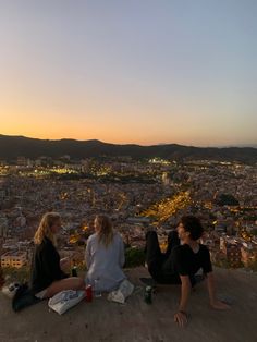 three people sitting on top of a hill overlooking a city at sunset with lights in the distance