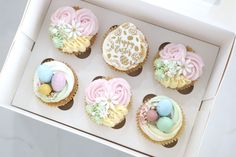 six decorated cupcakes in a white box on a marble countertop with eggs and flowers