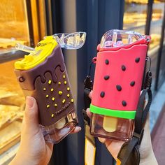 two ice cream sandwiches are being held up in front of a store window, one is shaped like a watermelon and the other is made out of plastic