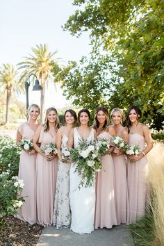 a group of women standing next to each other in front of trees and bushes holding bouquets