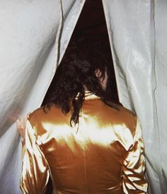 a woman with long hair standing in front of a white curtain and wearing a gold jacket