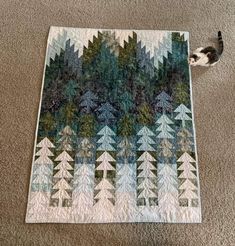 a cat is sitting on the floor next to a quilt that has trees in it