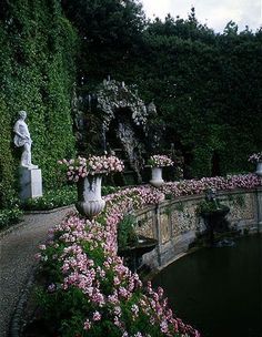 a garden with flowers and statues in the center, along side a pond surrounded by greenery