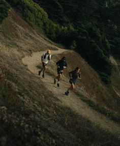 three people running down a trail in the woods on a hill side with trees and bushes behind them