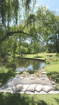 an outdoor table set up with pillows and flowers on the grass next to a river