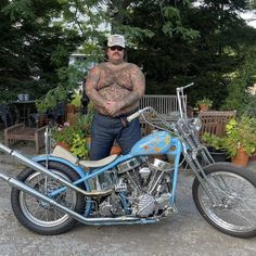 a man standing next to a blue motorcycle in a driveway with potted plants behind him