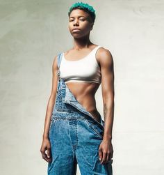 Photography by Studio PI artist, Kofi Paintsil of model Amosu M for Levi’s.   A ¾ shot of model Amosu. She is a young black female with a high top fade that has green tips. She is wearing a white sports bra and blue denim dungarees revealing her mid reefs and tattoo on her left arm. Photography Fashion, Overall Shorts