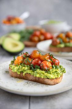 an avocado toast with tomatoes and other toppings on a white plate next to sliced avocado