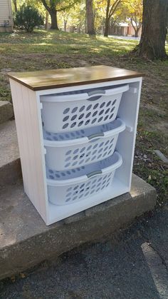 a white cabinet with three baskets in it on concrete steps next to grass and trees