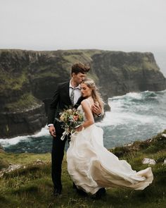 a bride and groom standing on top of a cliff overlooking the ocean with cliffs in the background