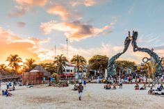 people are sitting on the beach and walking around in front of a large metal sculpture