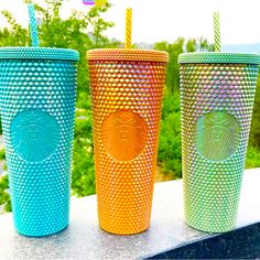 three different colored starbucks cups sitting next to each other on a table with trees in the background