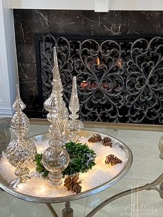 a glass table with candles and pine cones on it in front of a fire place