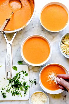 several bowls of soup are being prepared on a marble table with silver spoons and parmesan cheese