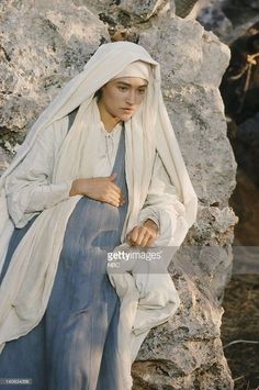 a woman dressed in white sitting next to some rocks and looking at the camera with her hand on her hip