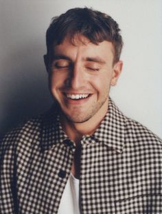 a man smiling and wearing a checkered shirt with his eyes closed, in front of a white wall