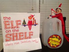 an elf is sitting next to a sign and a plastic jug that says the elf on the shelf