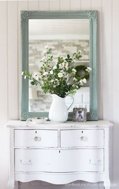 a white dresser topped with a vase filled with flowers next to a mirror and frame