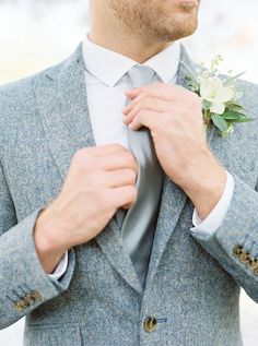 a man adjusting his tie while wearing a gray suit and white flower boutonniere