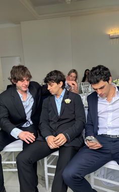 three men in suits sitting next to each other at a formal event with people standing around