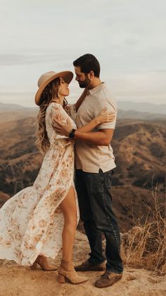 a man and woman standing on top of a mountain hugging each other with mountains in the background