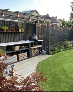 an outdoor kitchen and grill area with lights strung over the back yard, surrounded by greenery