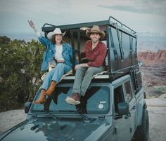 two people sitting on the roof of a vehicle with their arms in the air and smiling